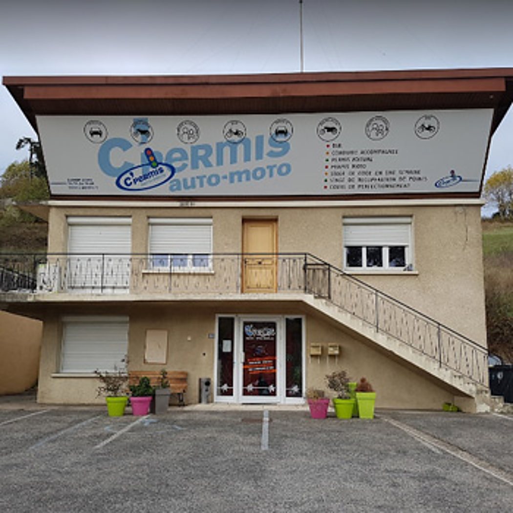 Façade d'un bâtiment de deux étages abritant une auto-école auto-moto, avec une grande enseigne, un escalier extérieur, des pots de fleurs colorés et un parking devant.