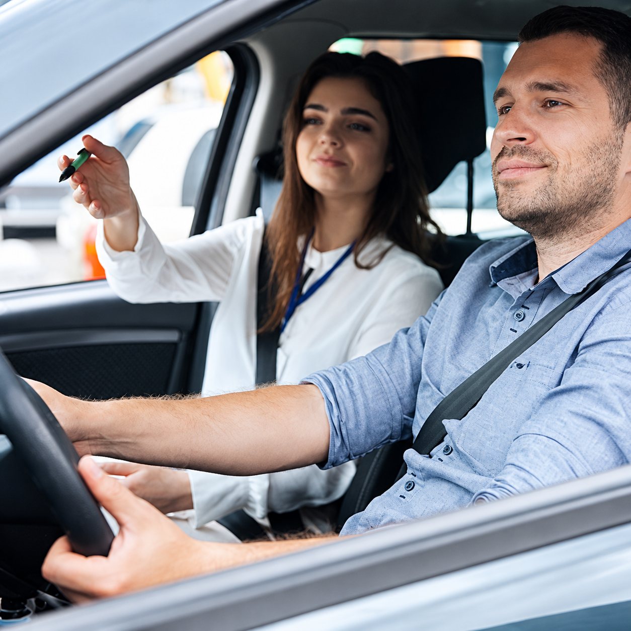 Un homme au volant d'une voiture écoute les consignes d'une monitrice assise à côté de lui, qui tient un stylo et gesticule.