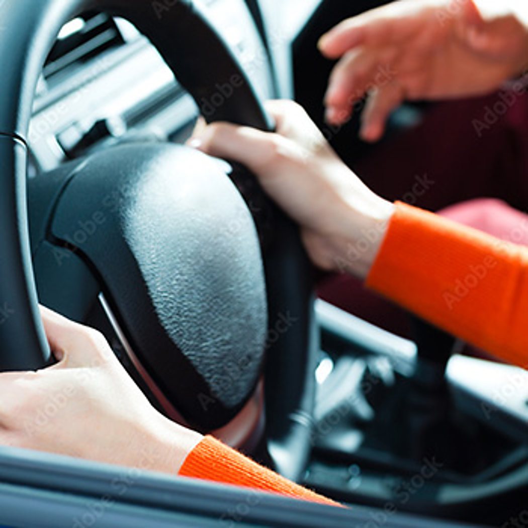 Deux personnes aux manches orange sont dans une voiture, l'une tenant le volant tandis que l'autre pointe la main vers le tableau de bord pendant une leçon de conduite.