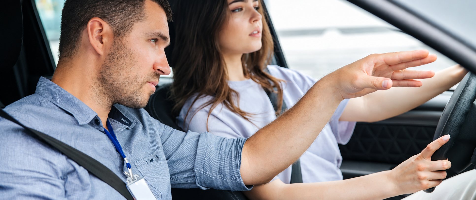 Dans une voiture, un moniteur en chemise bleue tend le bras pour guider une jeune conductrice qui tient le volant pendant une leçon de conduite.