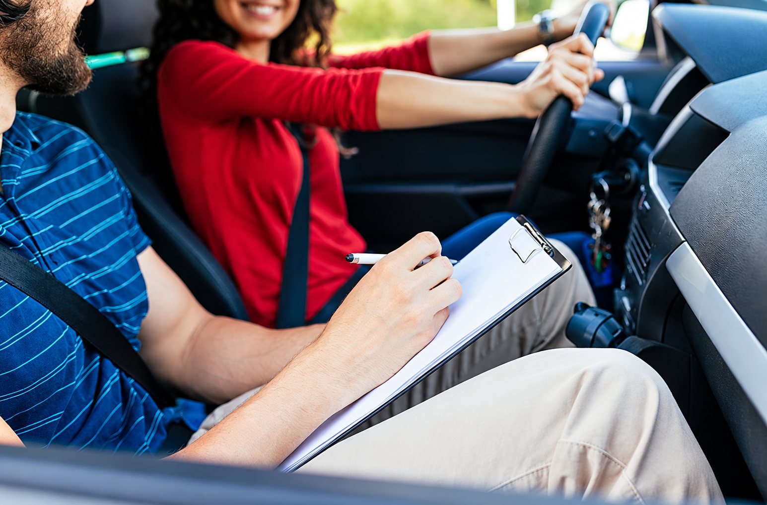 Dans une voiture, un moniteur barbu prend des notes sur un presse-papiers pendant qu'une jeune femme souriante en rouge tient le volant.
