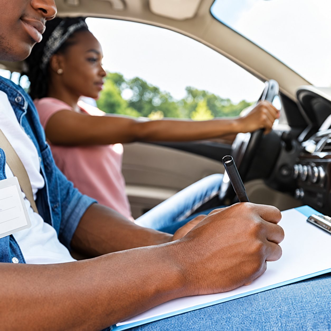 Dans une voiture, un moniteur écrit sur un bloc-notes tandis qu'une jeune femme au volant suit sa leçon de conduite.