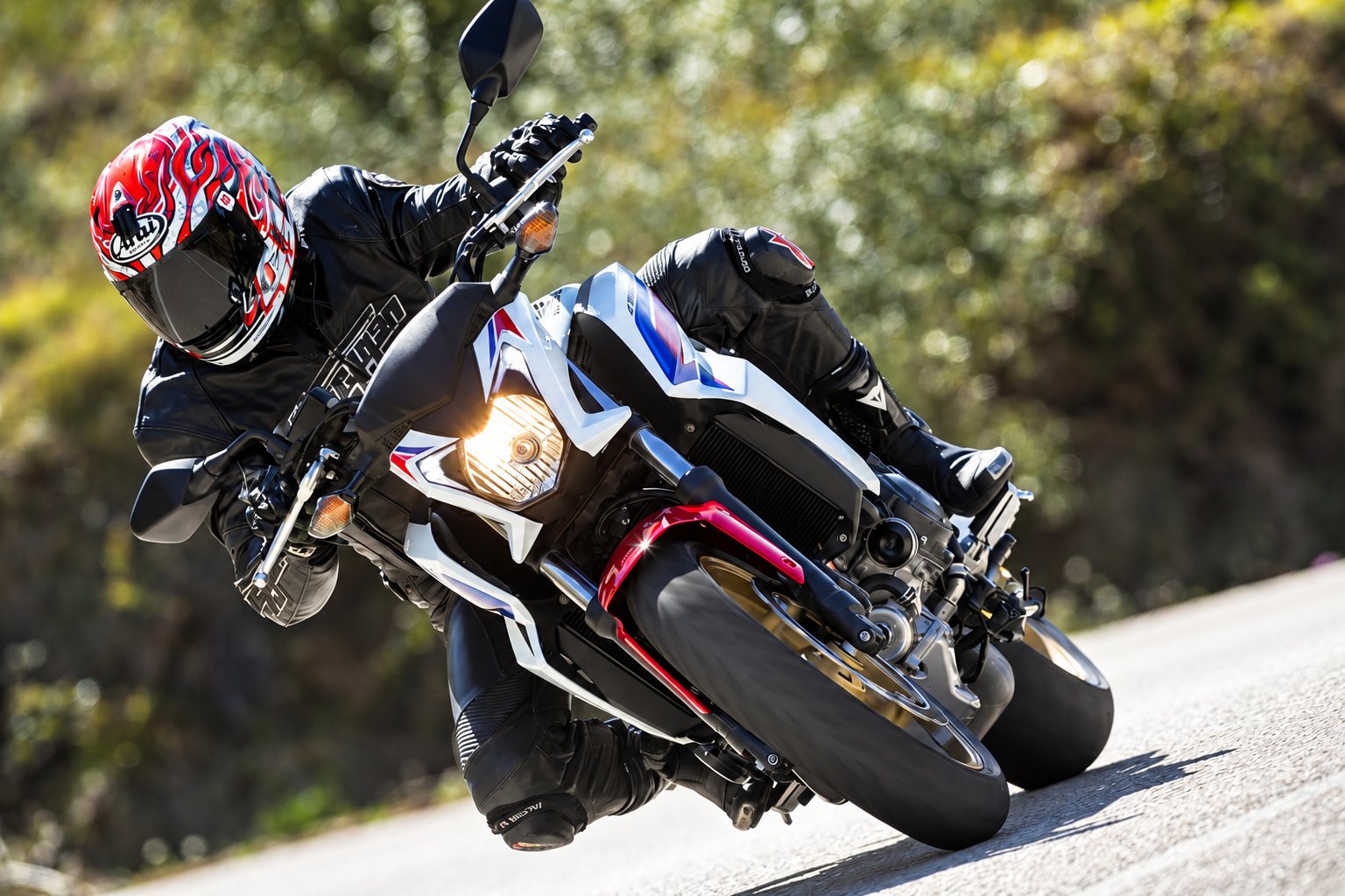 Un motard casqué en tenue de cuir noir incline sa moto sportive blanche, rouge et bleue dans un virage sur une route ensoleillée.