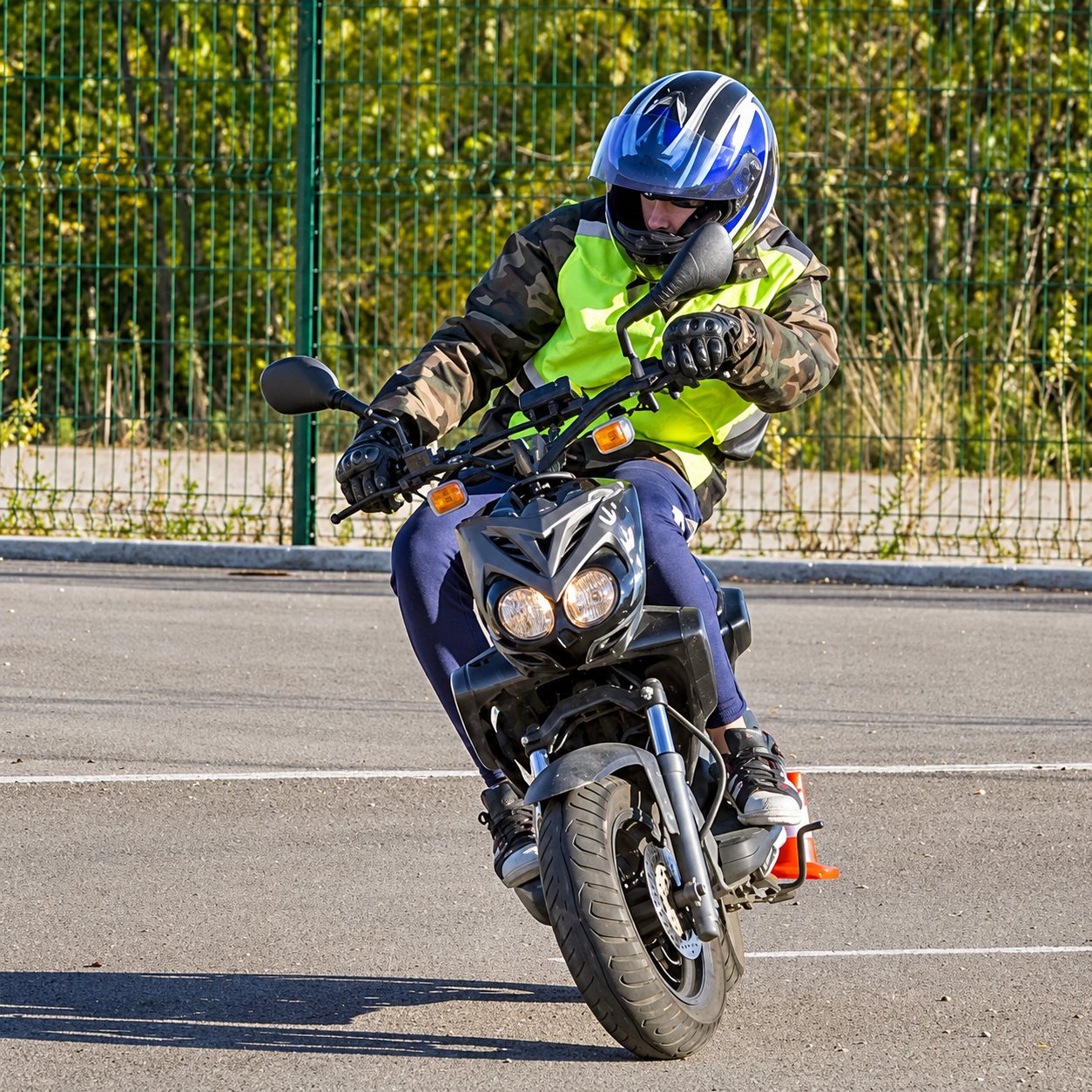 Un motard casqué en gilet jaune fluo incline un scooter noir pendant un exercice de maniabilité sur un terrain goudronné clos par un grillage.