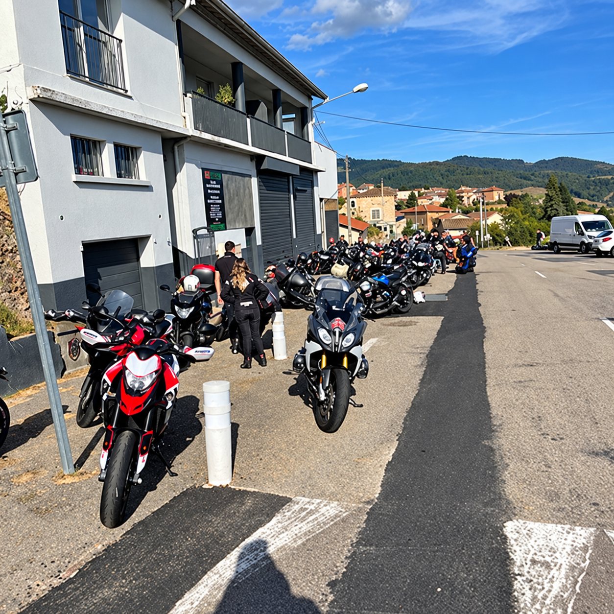 De nombreuses motos sont garées le long d'une route devant un bâtiment gris, avec des personnes debout et des collines en arrière-plan.